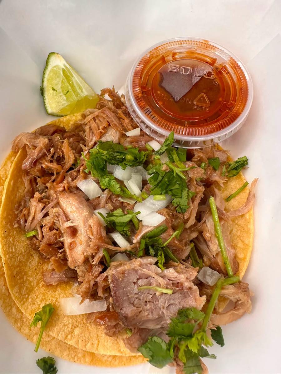 A carnitas street taco on a double corn tortilla, topped with onion and cilantro, with a lime wedge and a cup of red salsa.